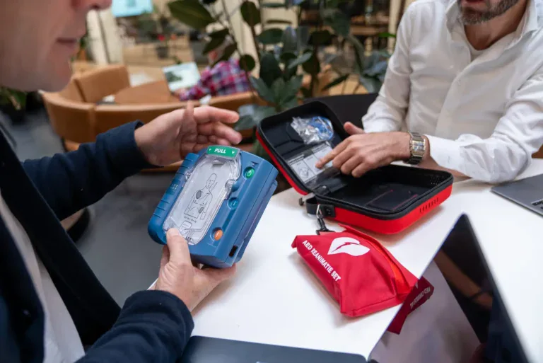 Twee mannen in gesprek aan een tafel met een geopende AED-set, waarbij een van hen een Philips AED vasthoudt en de ander naar de inhoud van de koffer wijst